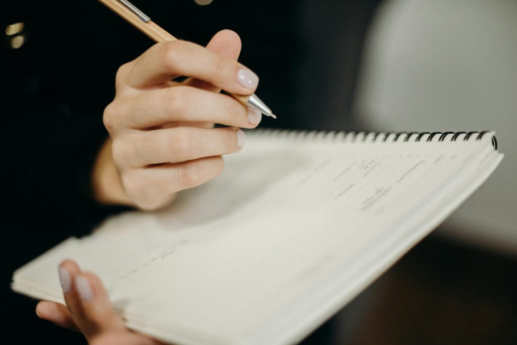 A person's hand holding a pen writing in a spiral notebook, focused on penmanship and note-taking.